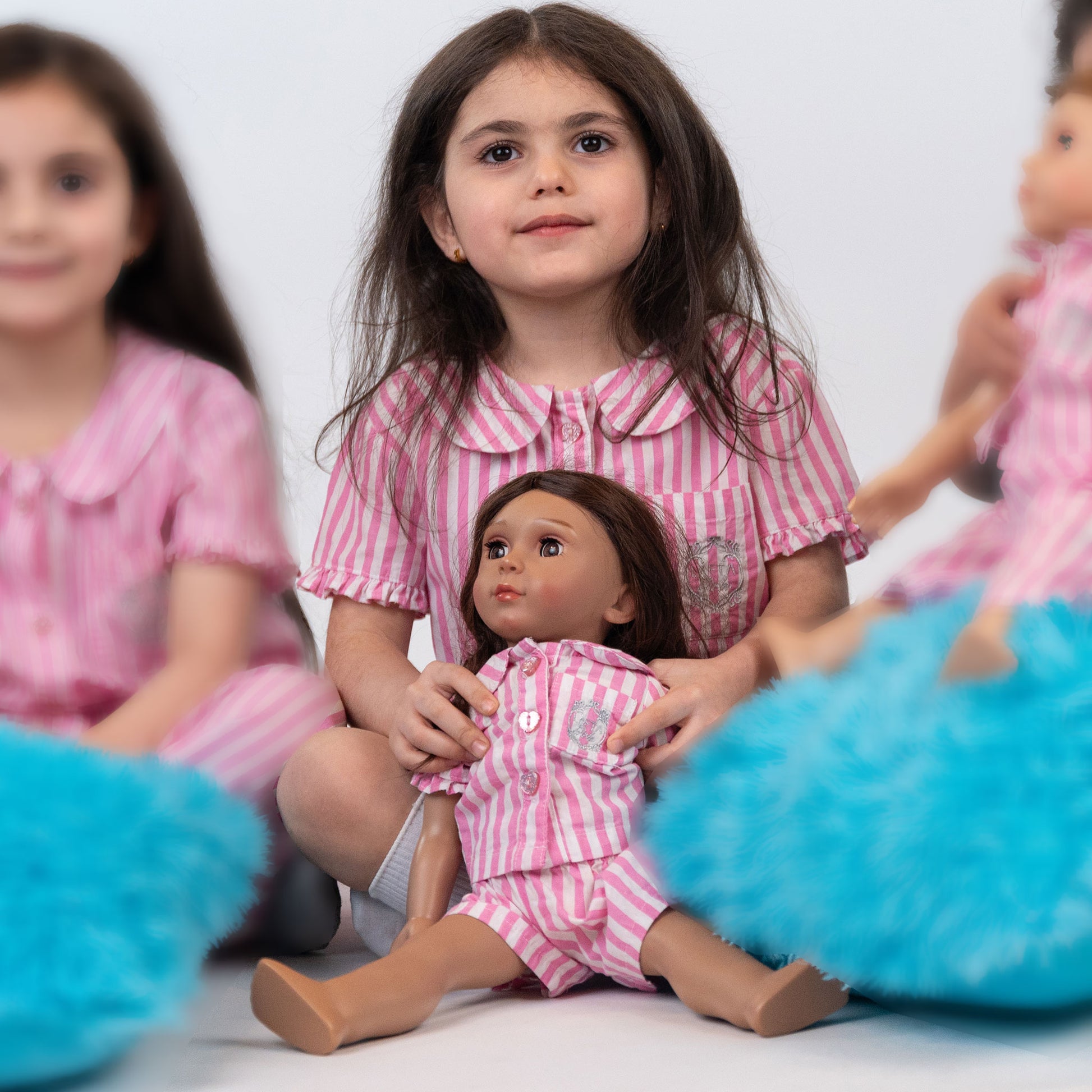 Picture of girl wearing LaftaKids short pajama sitting down and playing dress up with Yasmeen, the Little Me Doll, who is matching with the girl and wearing pink and white stripped short pajamas facing forwards