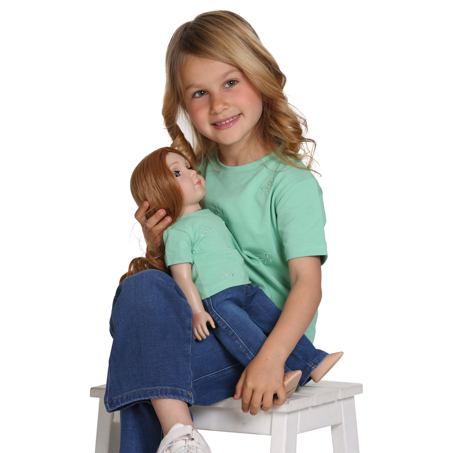 Girl wearing LaftaKids clothing of Casual green top and jeans holding a Little Me doll who is also wearing Casual green top and jeans, girl is sitting down and looking to the right