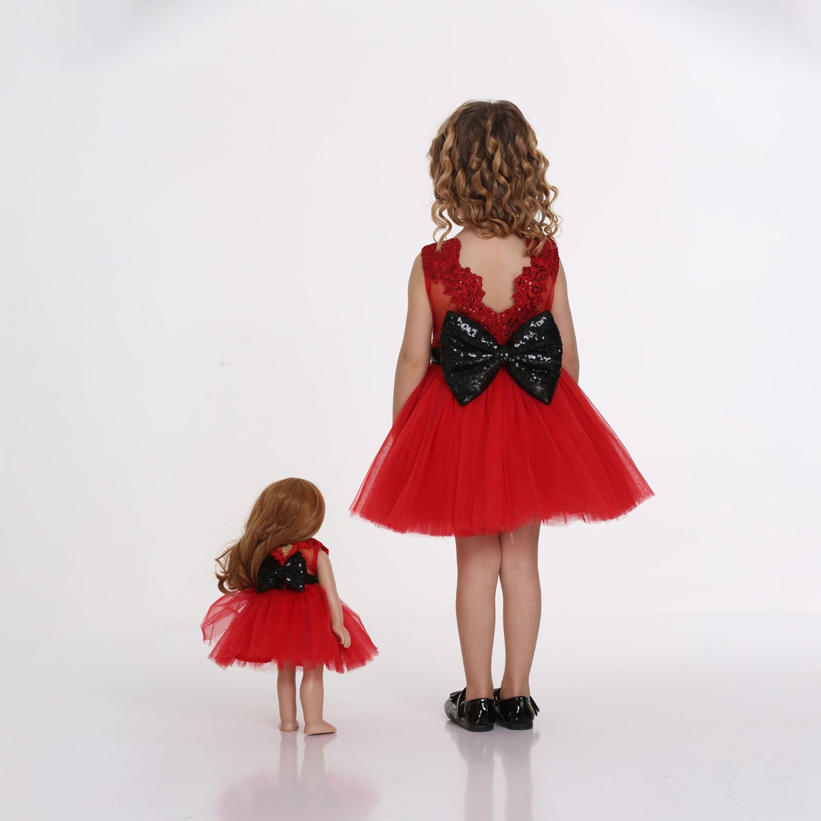 Girl Standing next to Mais, The Little Me Doll, both wearing red dress by laftakids, facing back to showcase the back of the dress which shows a big black sparkling bow