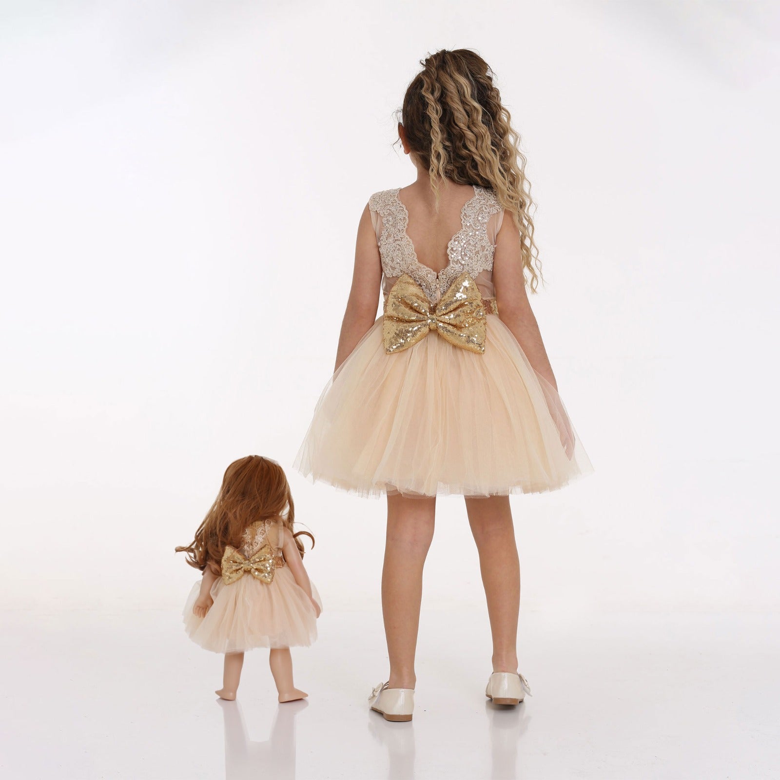 Girl and Mais, the Little Me Doll, wearing cream dress with sparkling gold belt facing backwards to showcase big gold bow at the back of dress