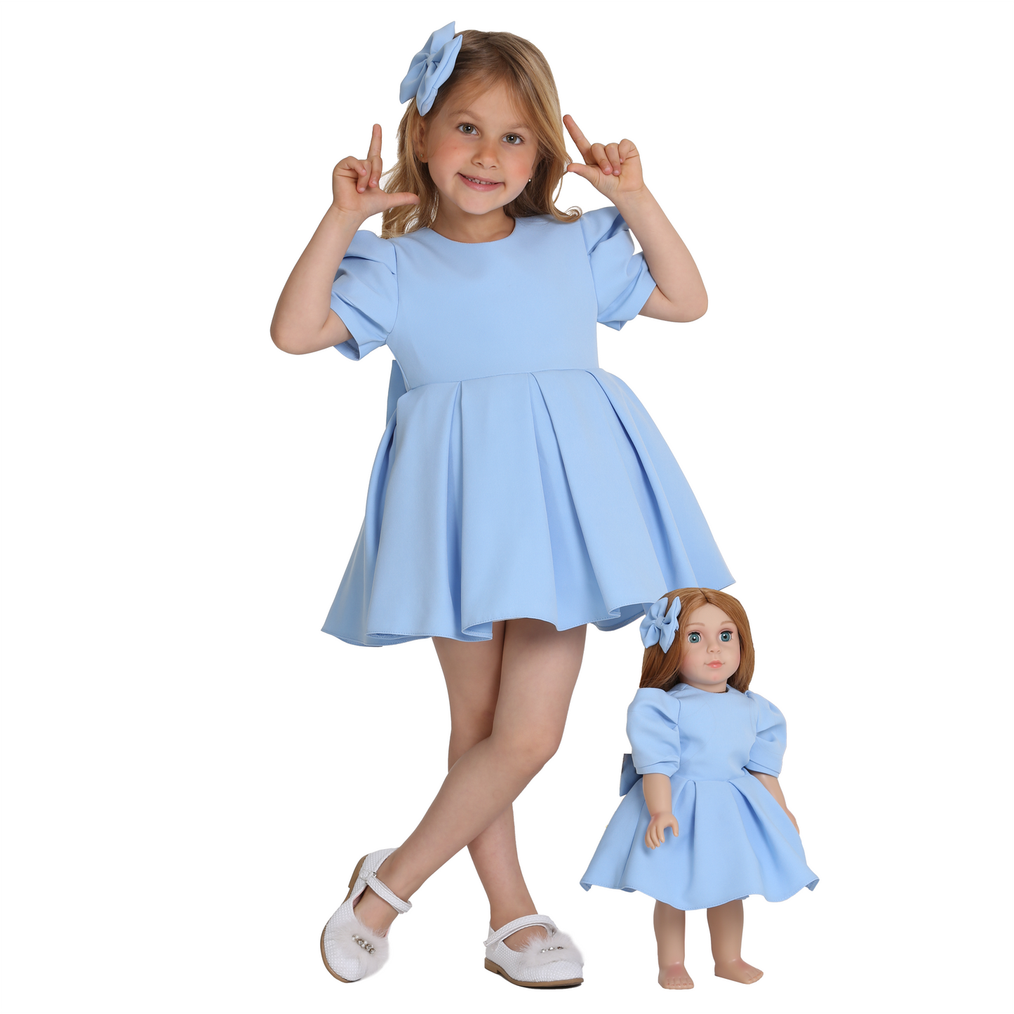 Girl wearing blue dress with a bow tie in hair, standing next to her doll with matching outfit, facing forward